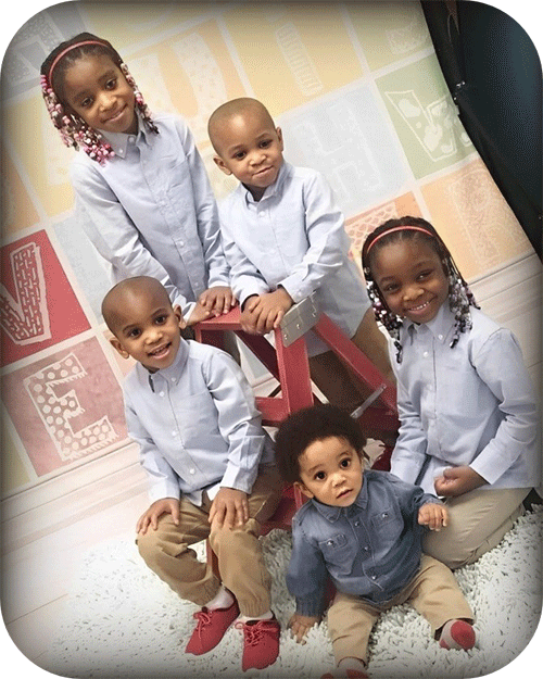 Five children, smiling, pose in a studio. They wear light blue shirts and khaki pants. One sits on a red stool.