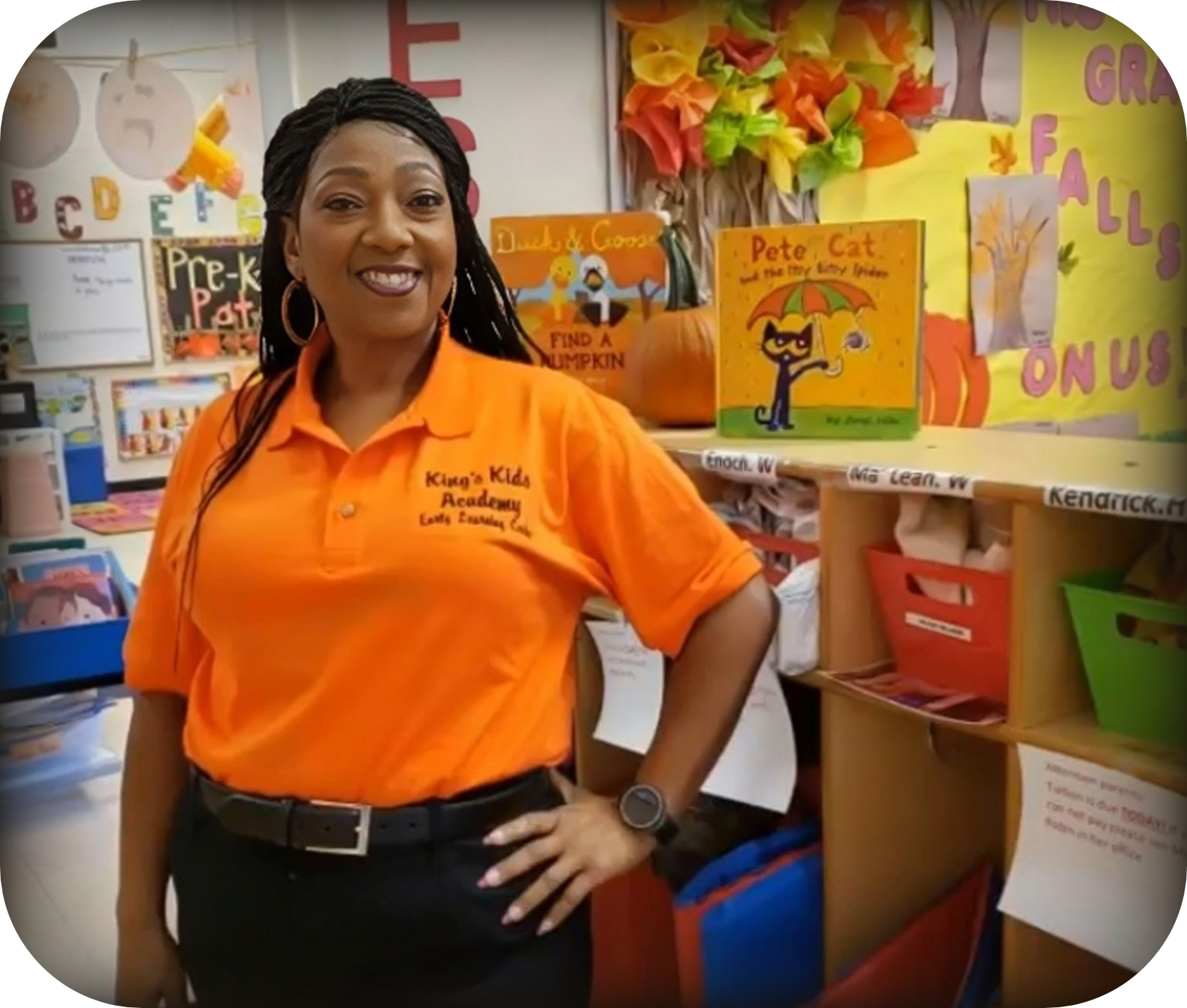 Woman in orange shirt, braids, smiling, in a classroom. Books and fall decorations are visible.