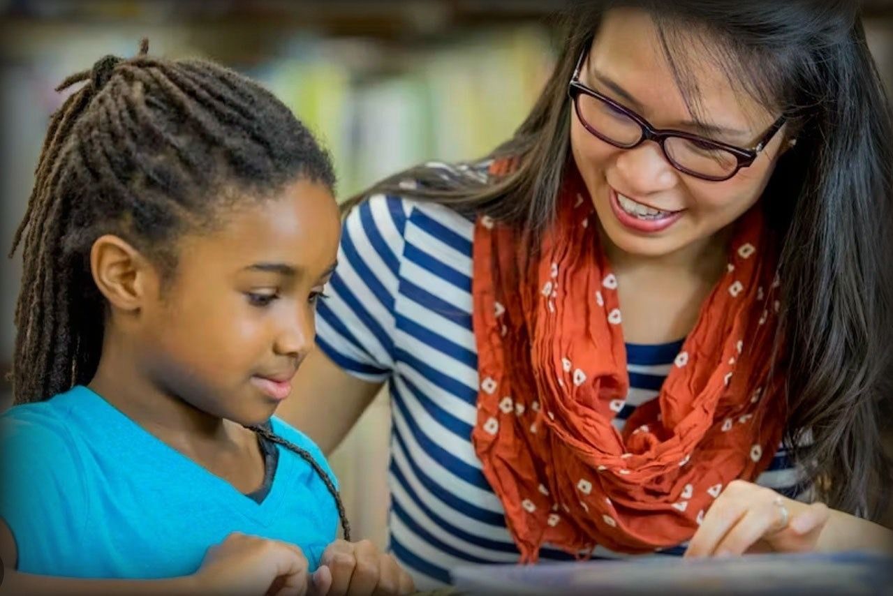 Teacher with glasses smiles, reads with child with dreadlocks in a library.
