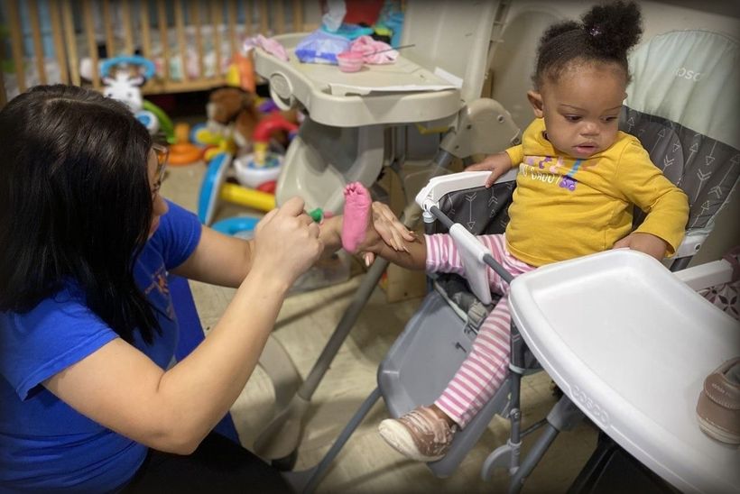 Woman in blue shirt trims a toddler's nails while she sits in a high chair.