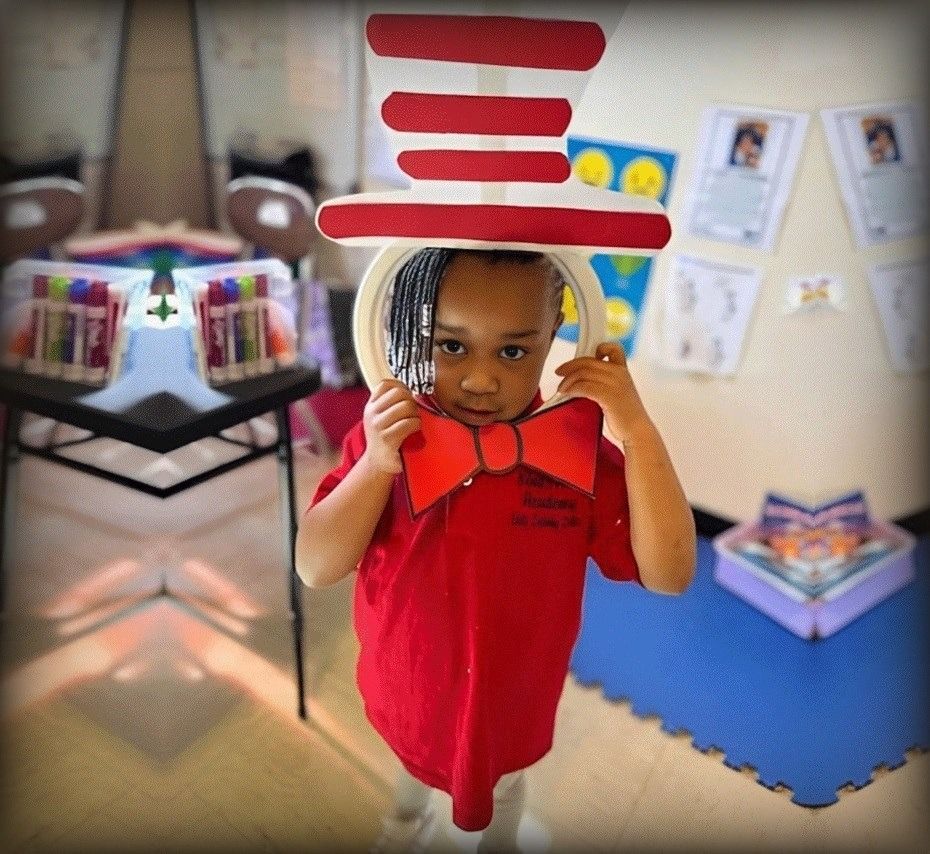 Child dressed as the Cat in the Hat, wearing a red shirt, bow tie, and hat, in a classroom.