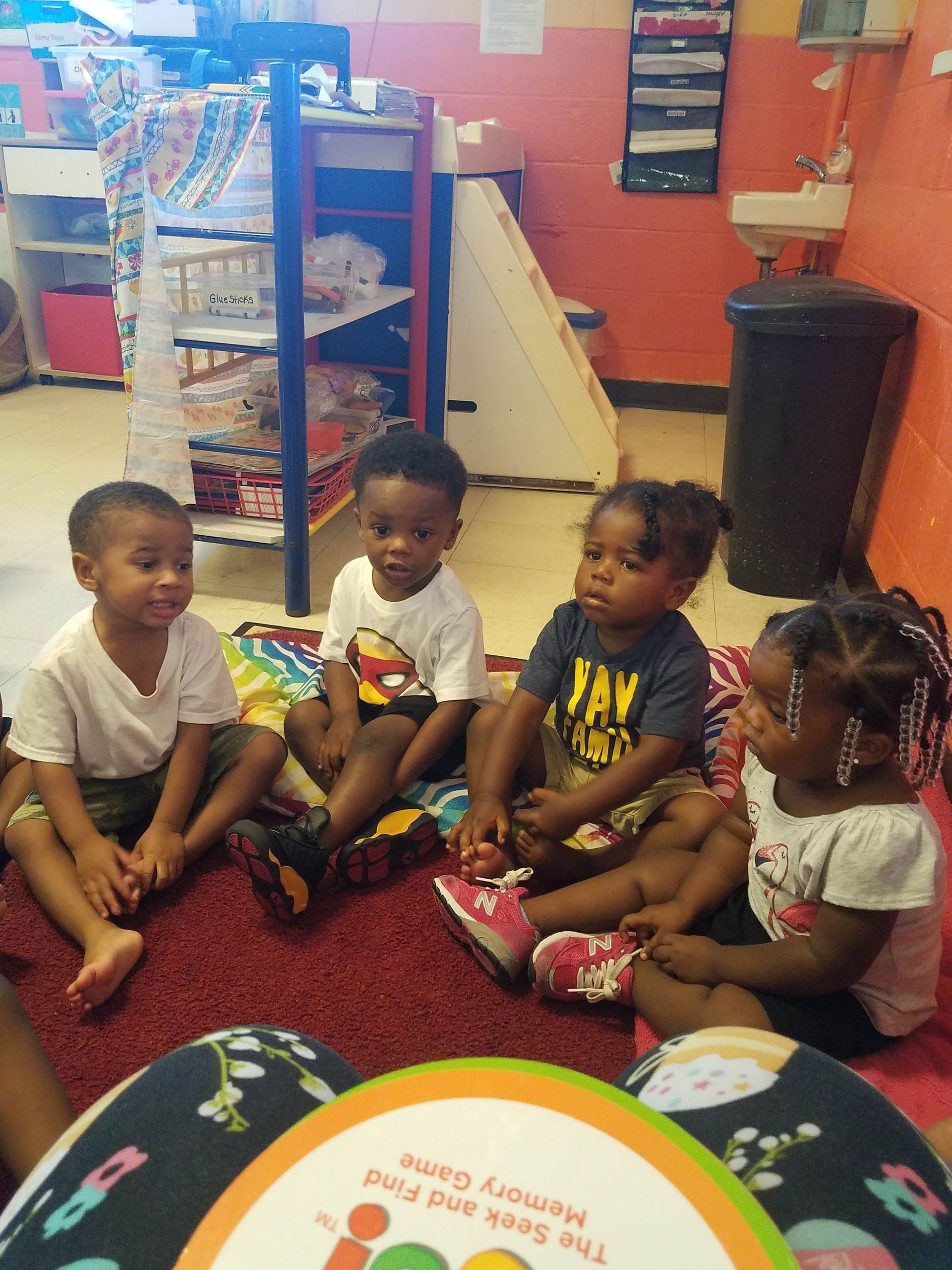 Four young children sit on a rug in a classroom, looking towards a person holding a game.
