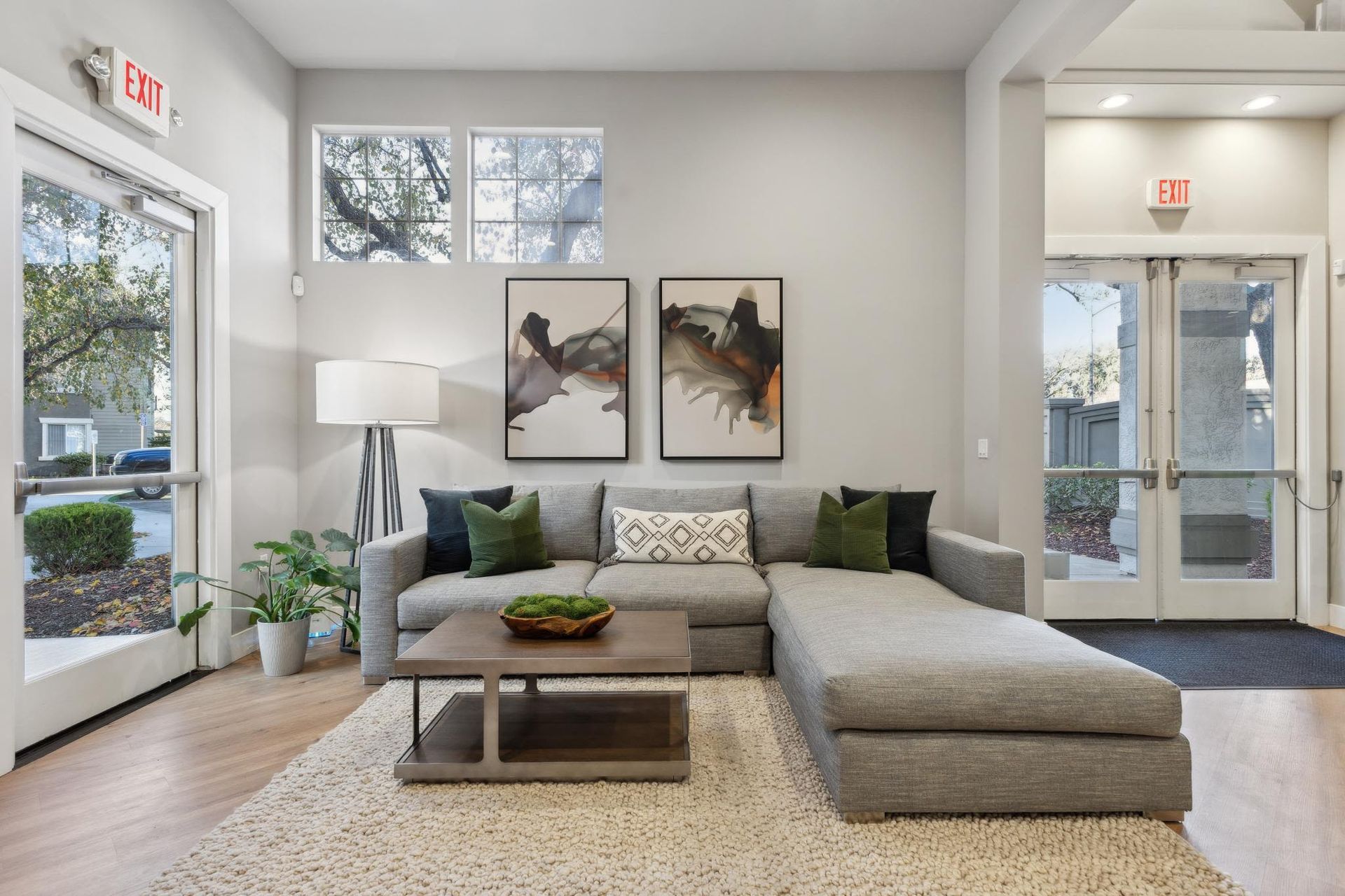 Living room in a modern apartment with a gray sectional sofa, glass coffee table, plants, and abstract wall art.