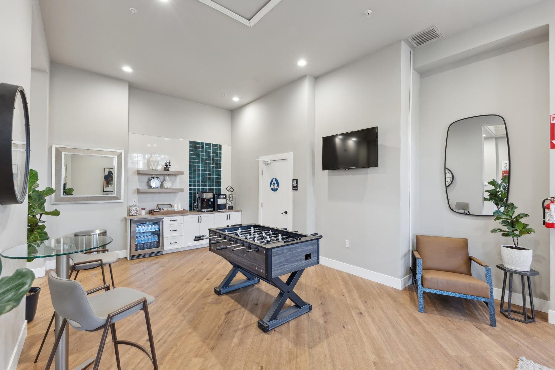 Common lounge with foosball table, kitchenette, wall TV, seating, and plants.