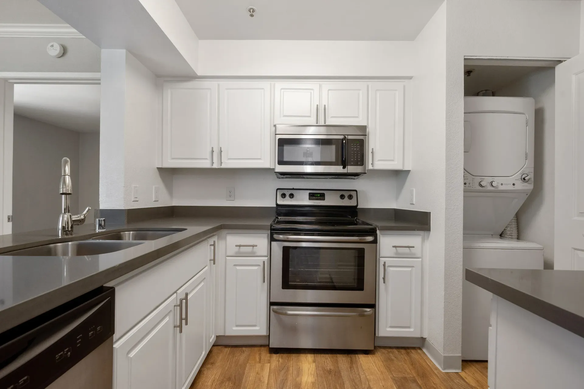 White kitchen with stainless steel stove, microwave, double sink, gray countertops, and a stacked washer/dryer in an adjacent alcove.