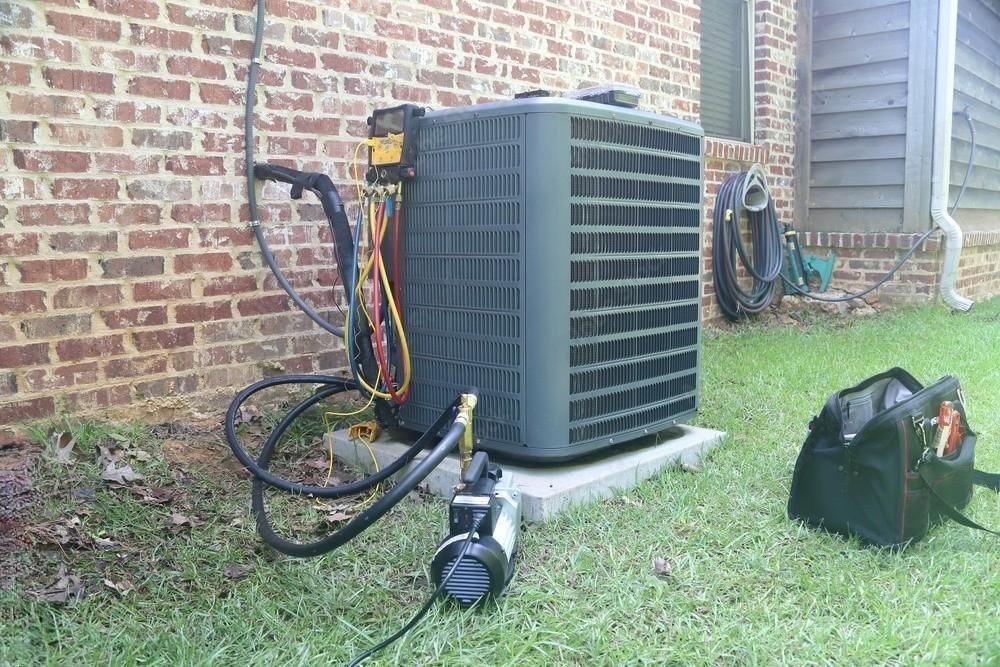 Outdoor HVAC unit beside a brick house with hoses, tools, and a black tool bag on the grass