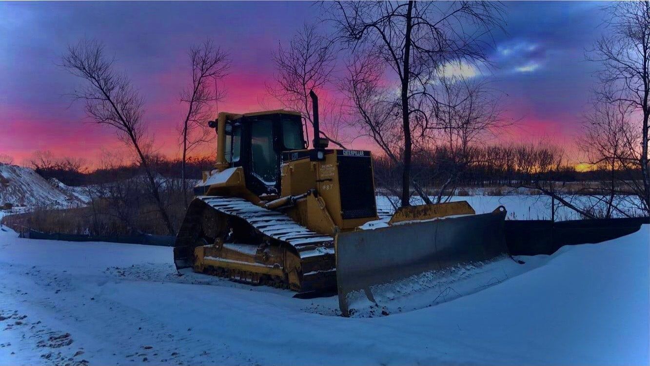 A bulldozer is parked in the snow on a snowy road at sunset.