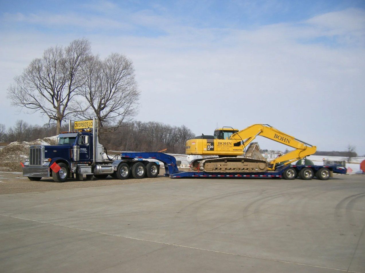 A blue semi truck is carrying a yellow excavator on a trailer