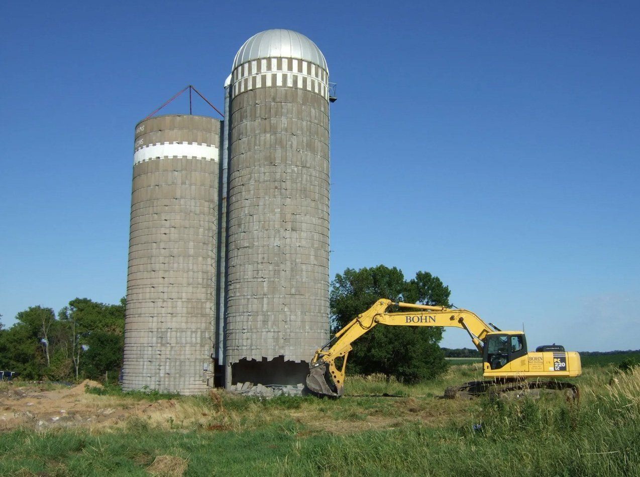 A yellow excavator is in front of two silos