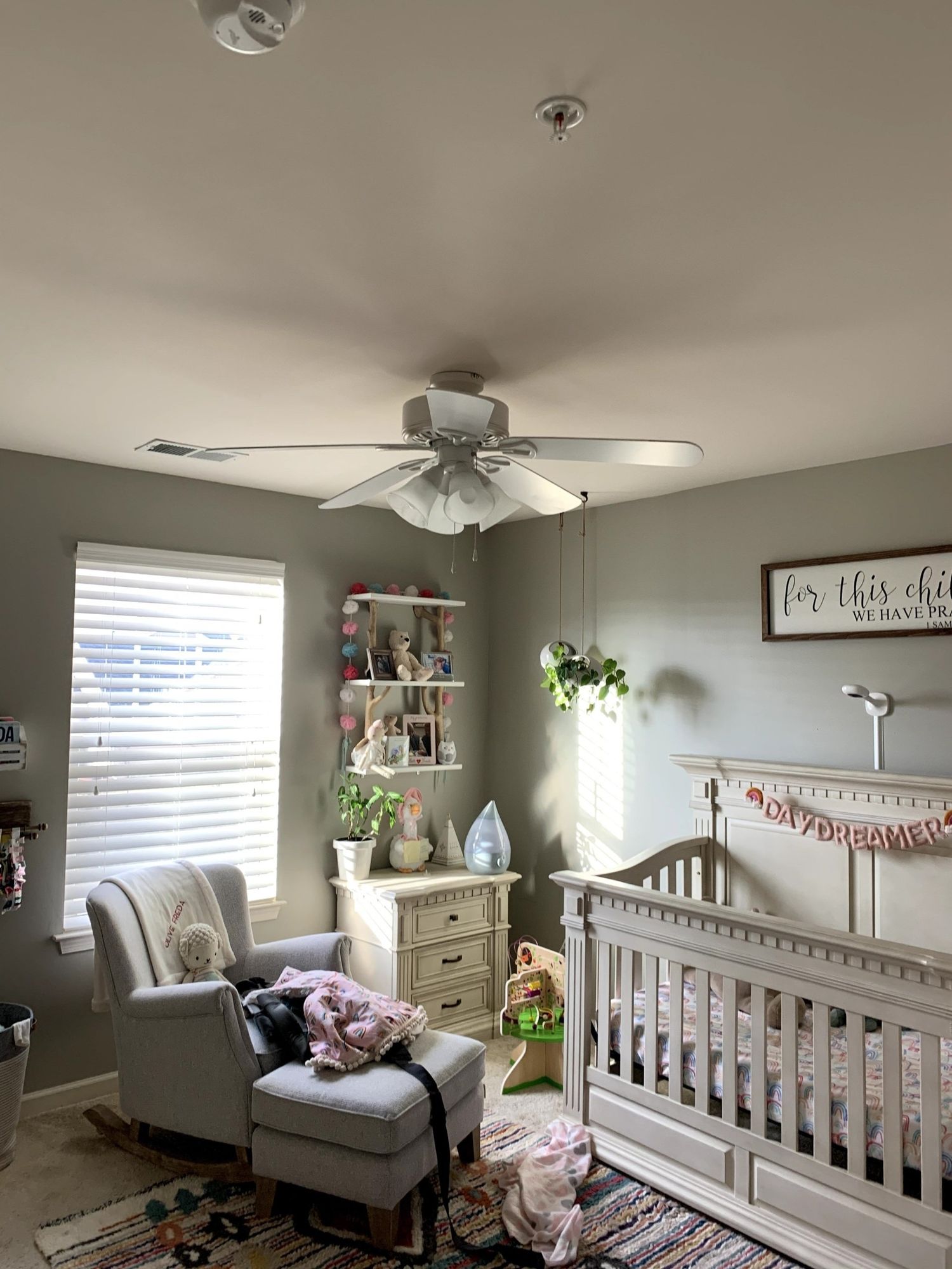 A baby room with a crib , rocking chair , ottoman and ceiling fan.