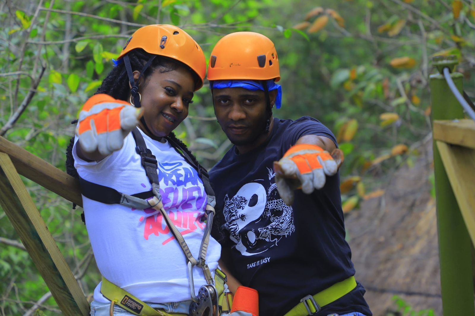 Couple in orange helmets, safety harnesses, and gloves. They point toward the camera, smiling, against a green forest background.