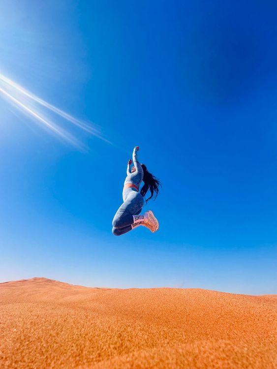 Woman jumping in the air against a bright blue sky, over a sandy desert landscape.