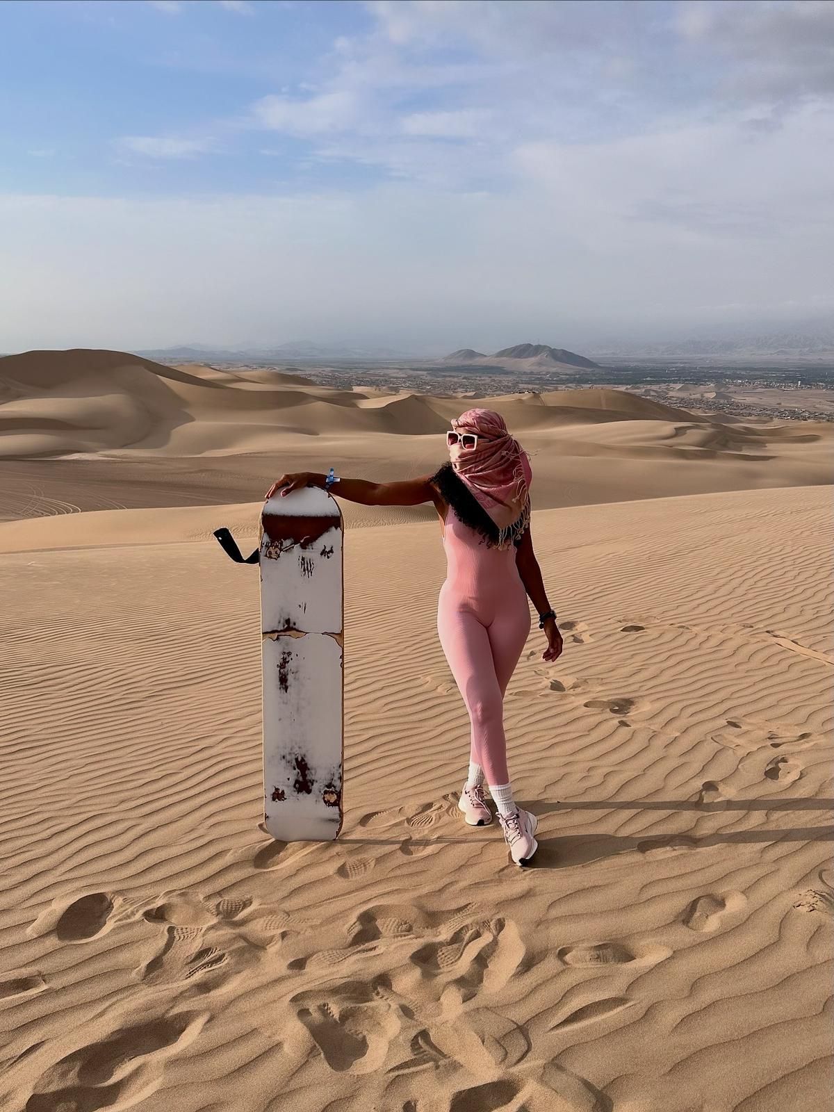 A person in a pink jumpsuit and headscarf leans on a white sandboard on a vast, rippled sand dune under a blue sky.