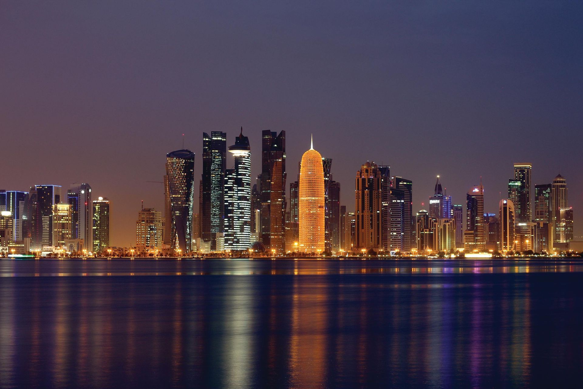 Doha, Qatar skyline at night, with illuminated skyscrapers reflected in calm water.