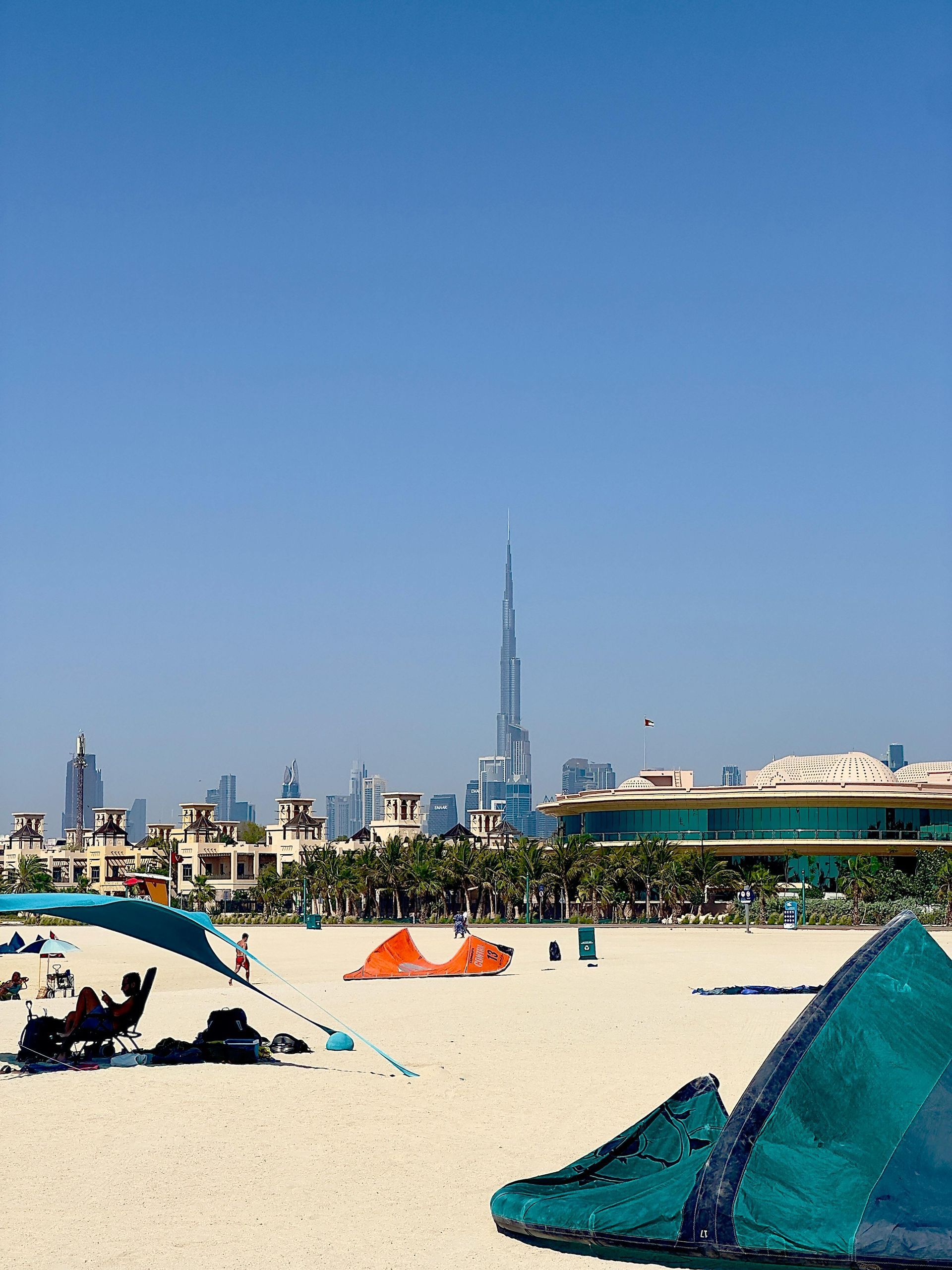 Beach scene in Dubai with Burj Khalifa skyscraper, beachgoers, tents, and blue sky.