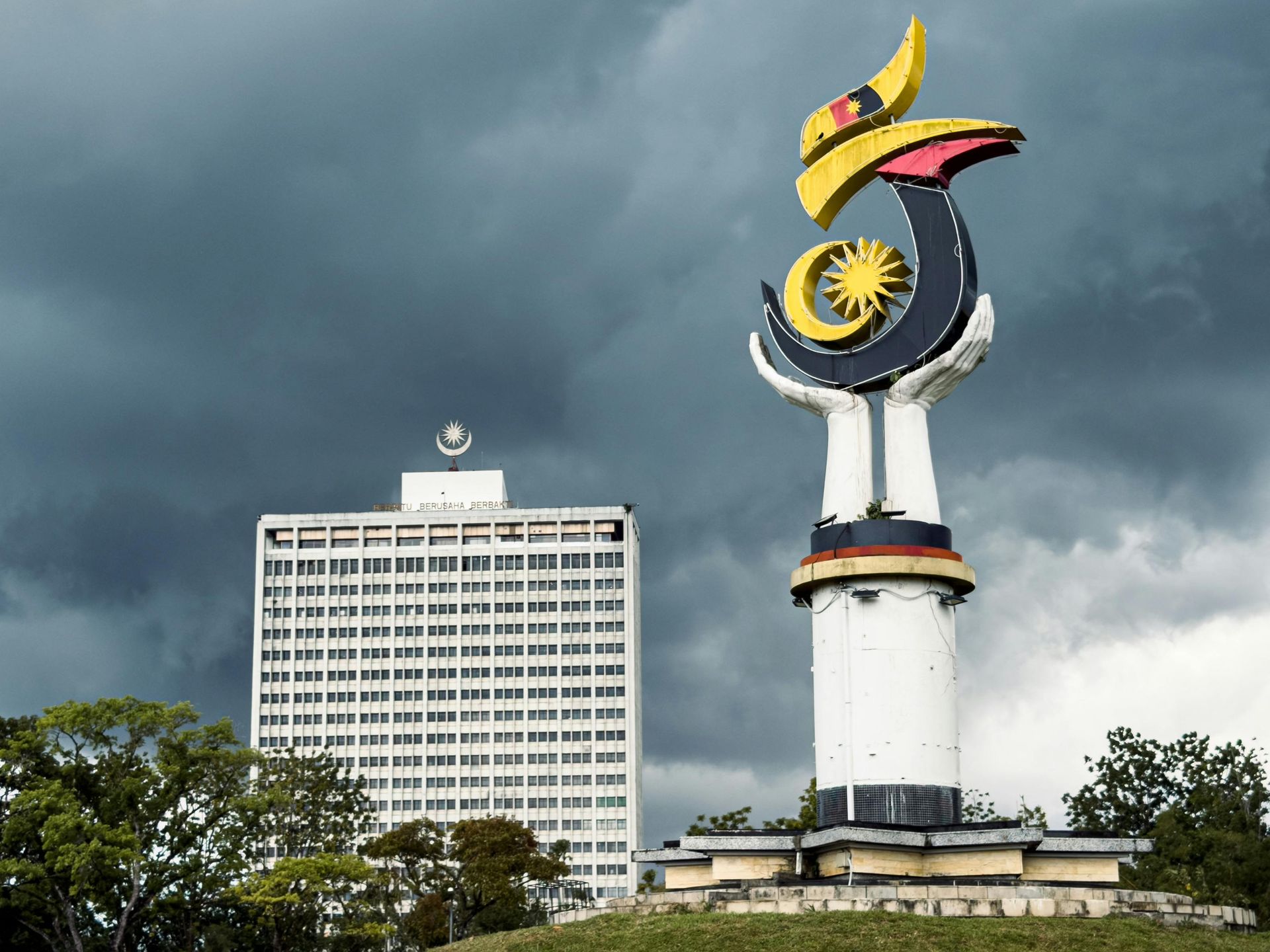 Monument with a hornbill and the Malaysian flag, and a tall white building under a stormy sky.
