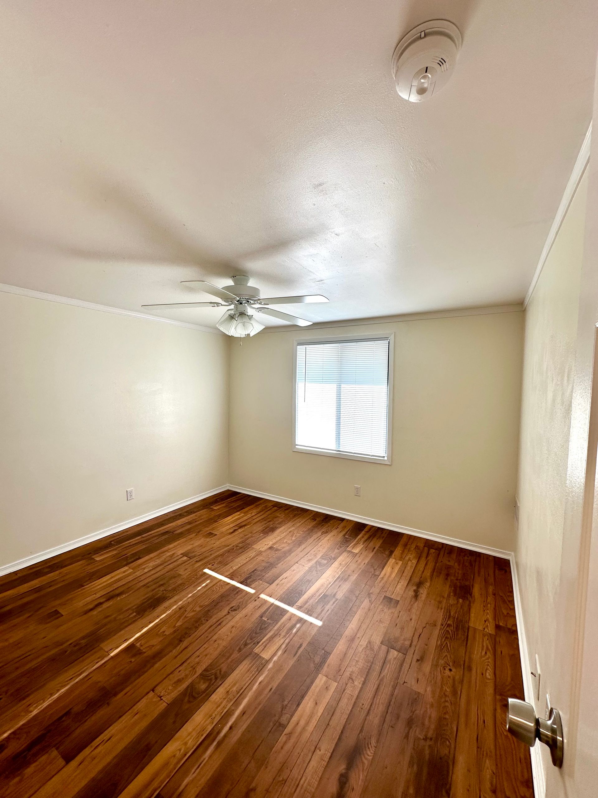 Empty room with hardwood floors, a ceiling fan, window with blinds, and a smoke detector.