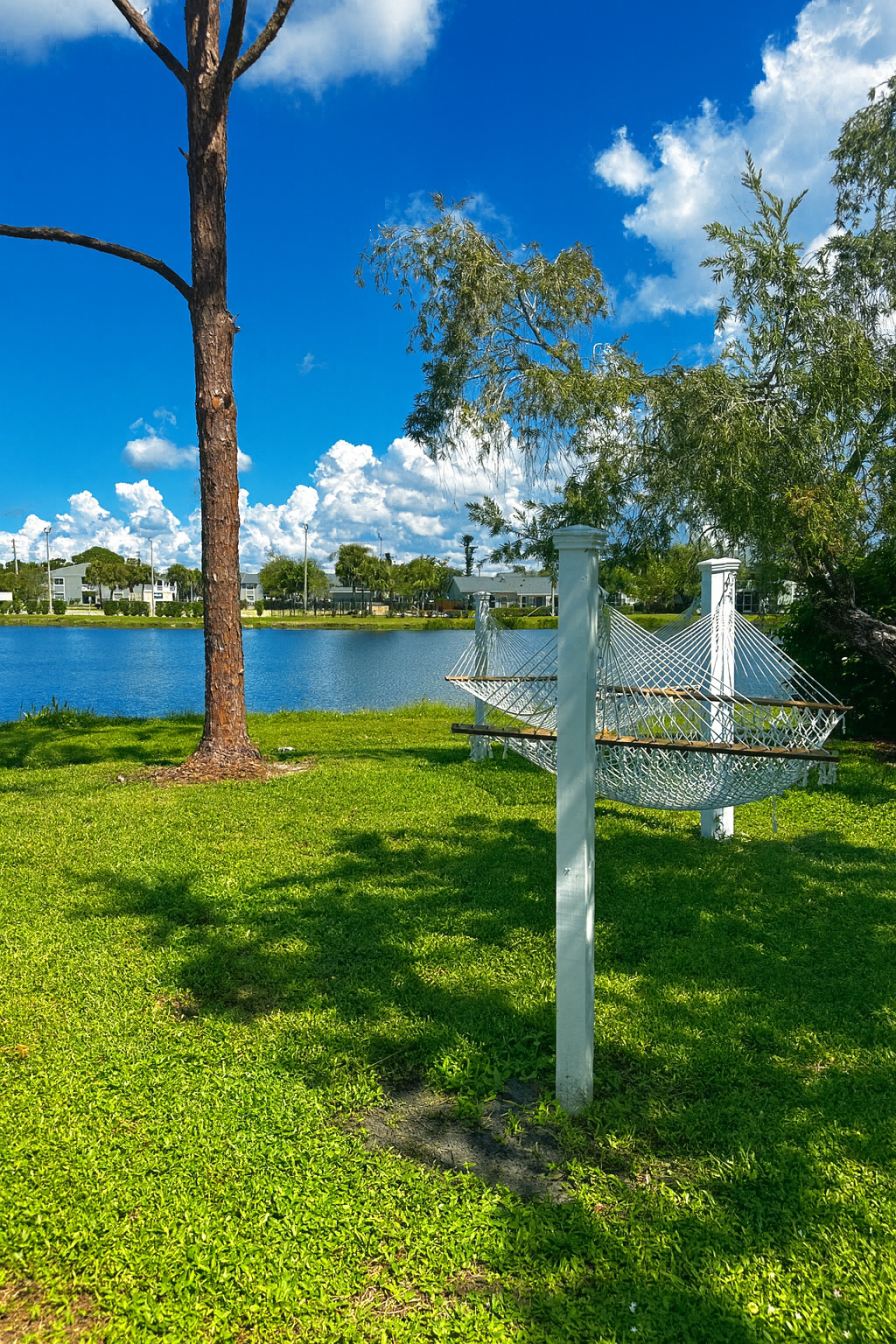 White hammock between two posts on grassy lawn by a lake under blue sky and clouds.