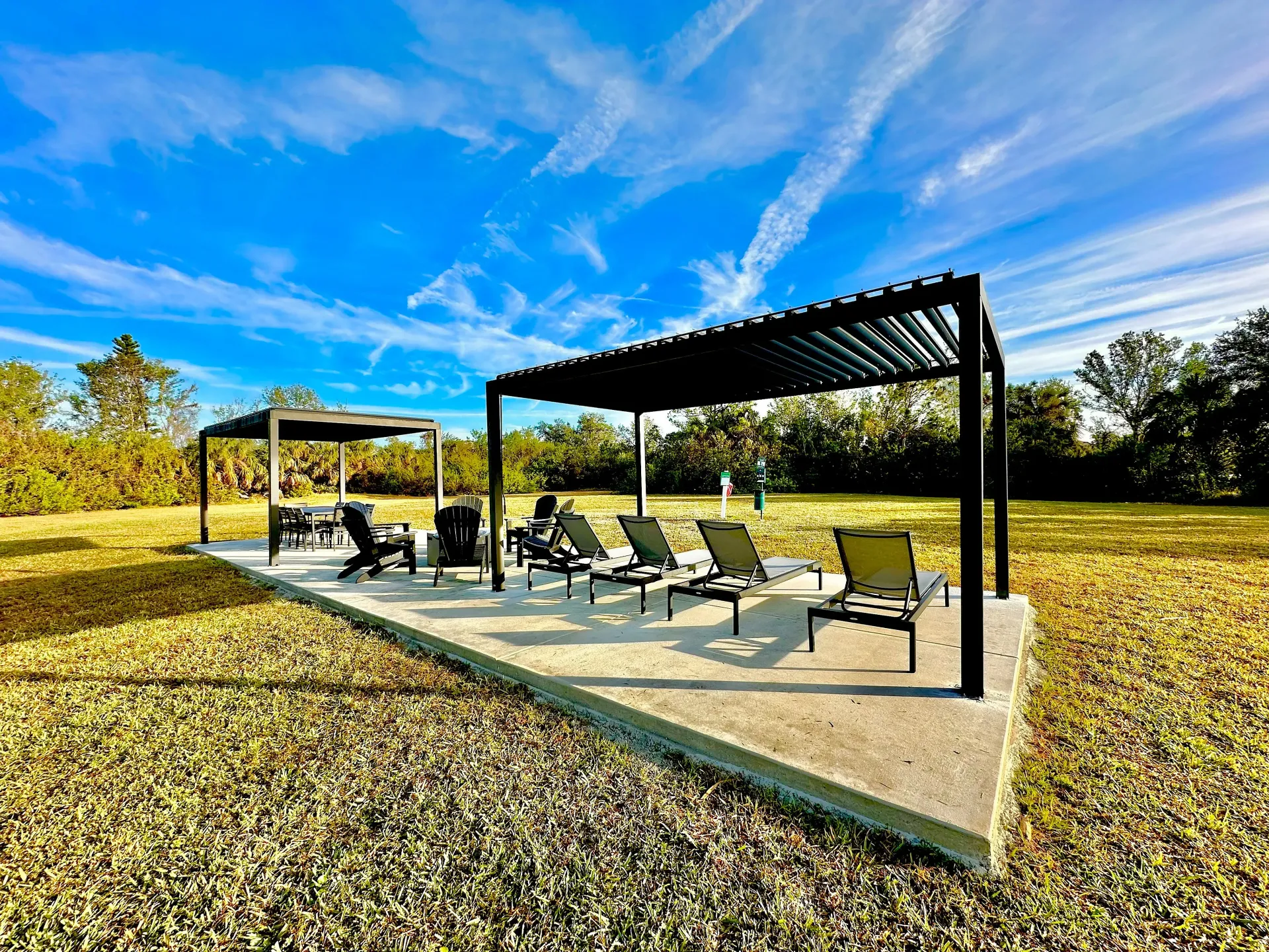 Two black pergolas with lounge chairs on concrete pad in grassy field under blue sky.