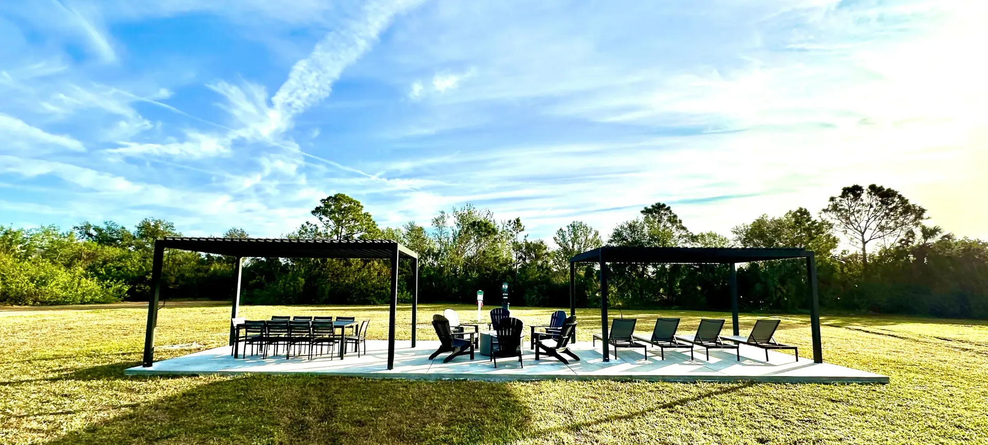 Outdoor seating area with pergolas on a concrete slab in a grassy field. Blue sky, trees in the background.
