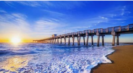 Ocean pier at sunrise, sun over the water, blue sky, waves on the beach.