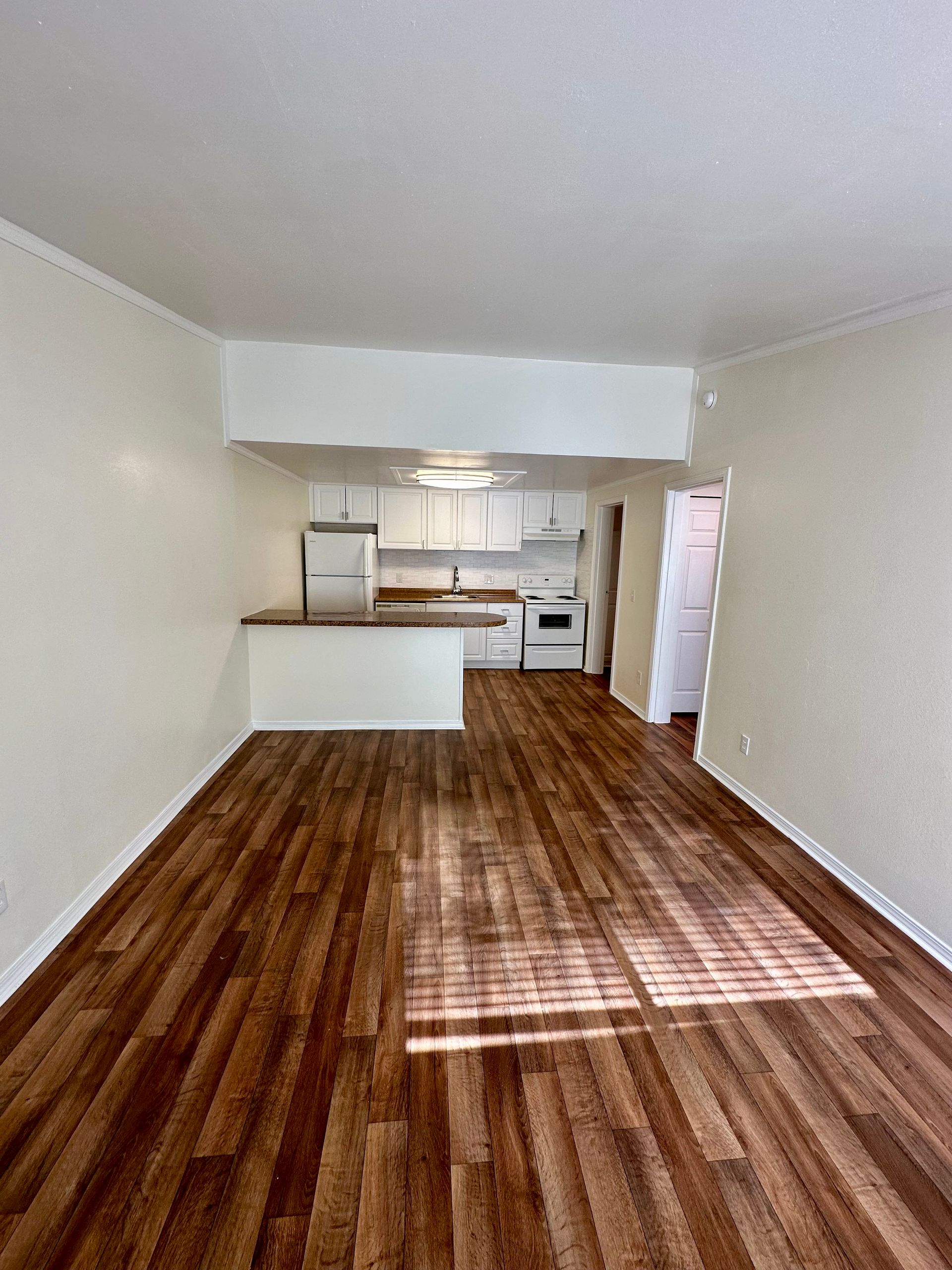 Empty living room with wood flooring, open to a white kitchen, and natural light.