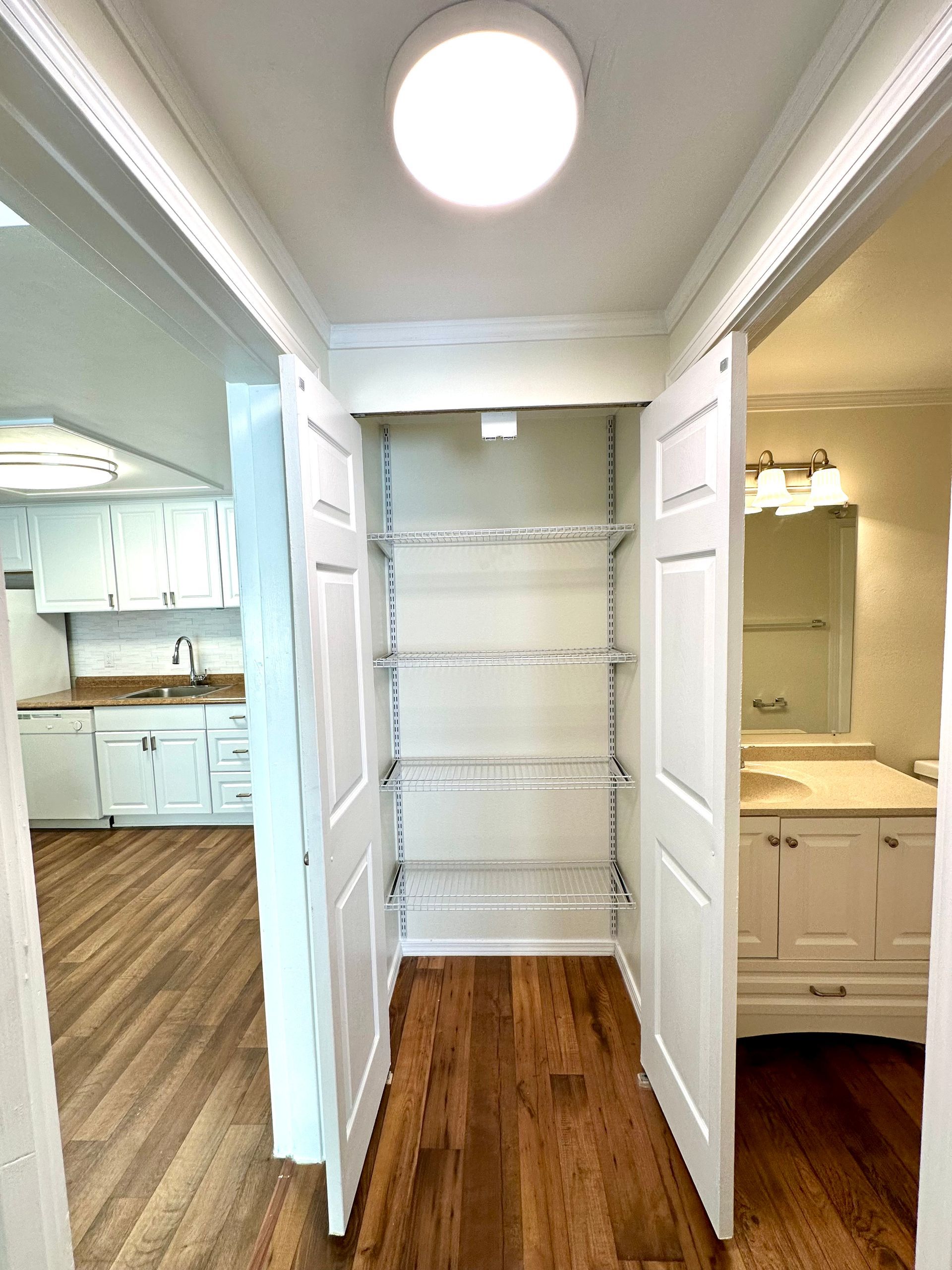 Narrow hallway with open white doors revealing wire shelving. Kitchen on left, bathroom on right.