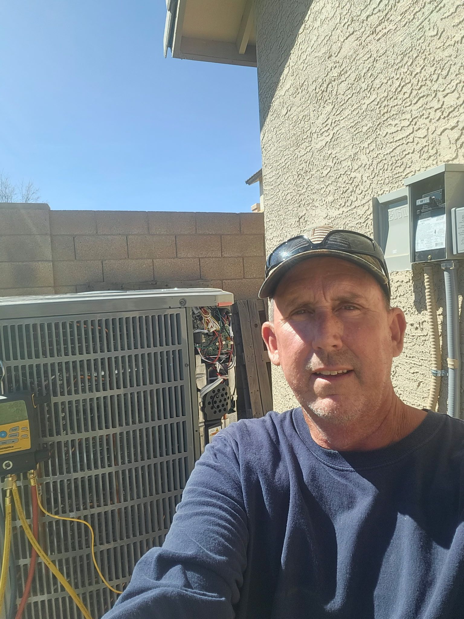 A man wearing a cap and blue shirt poses in front of an outdoor air conditioning unit next to a stucco house.