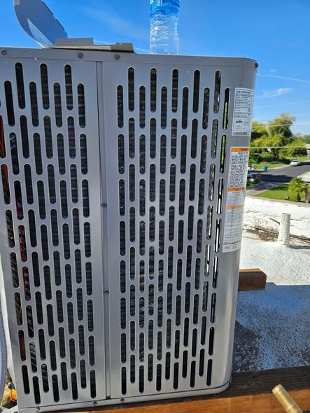 A close-up of a metal air conditioner condenser unit showing the textured, slatted side panel and a warning label.
