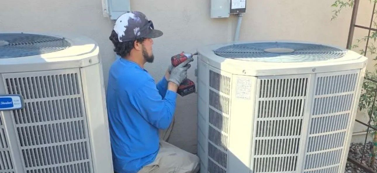 Technician kneels between two outdoor AC units, using a power drill on a wall-mounted connection