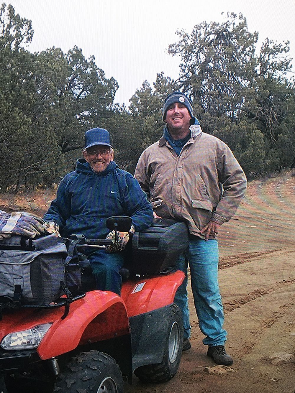 Two men are standing next to an atv on a dirt road.
