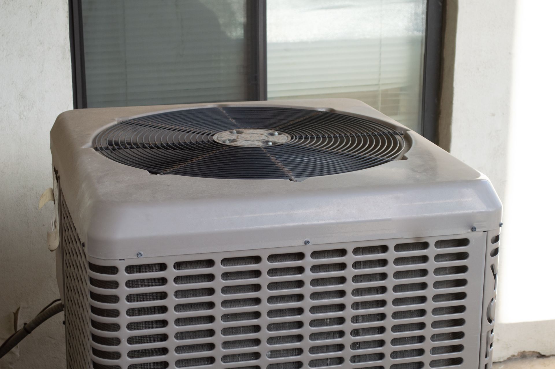 A light gray outdoor residential air conditioning unit sitting on a patio in front of a window.