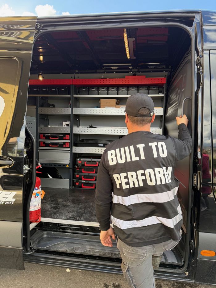 A man wearing a vest that says `` built to perform '' is standing in front of a van.
