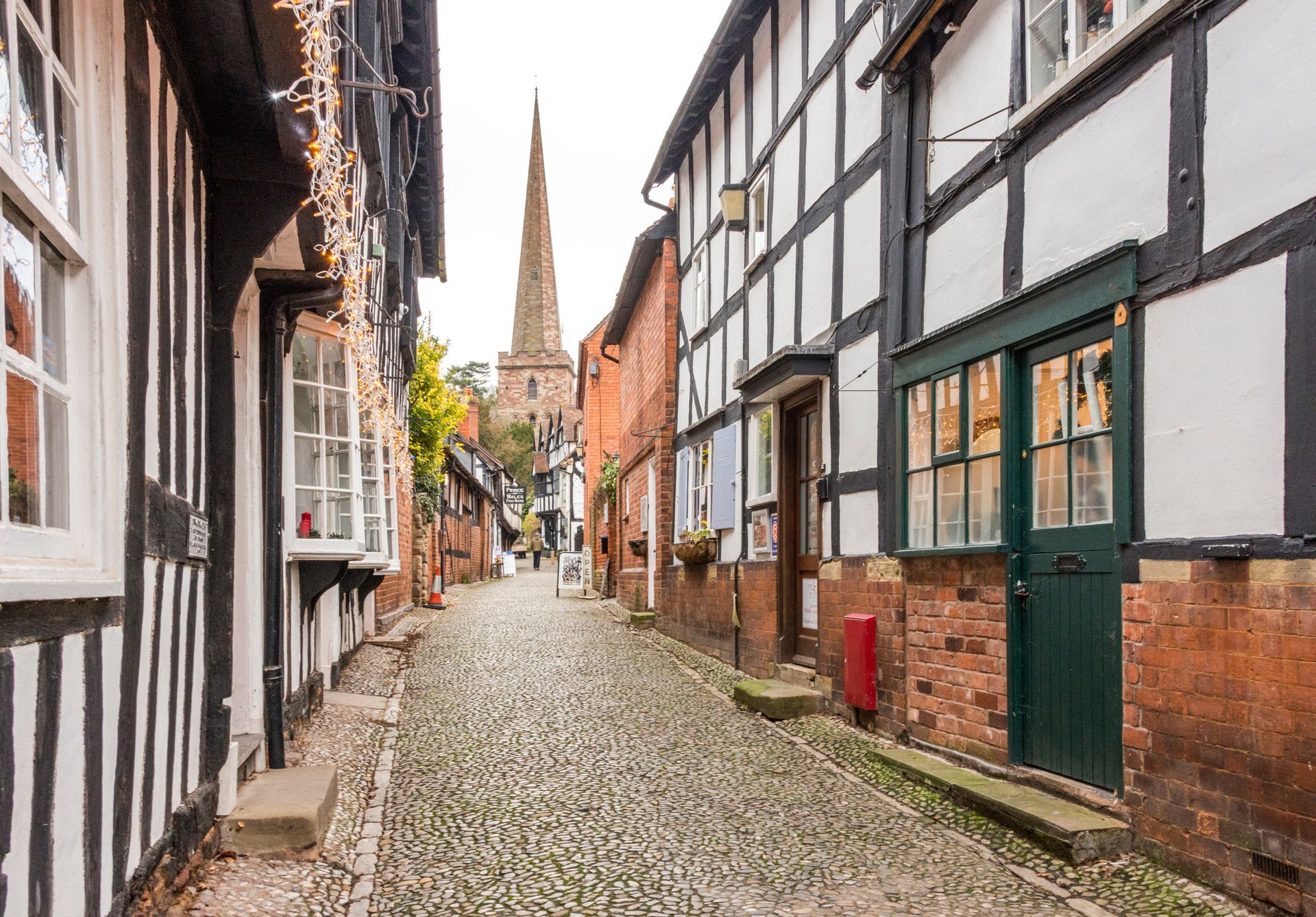 Photograph of a narrow and historic street in Ledbury, Herefordshire, England, United Kingdom. The houses are a mix of black and white Tudor circa 1485 - 1603 period properties and Victorian. Shown in the back ground is the Ledbury Church with a narrow steeple.