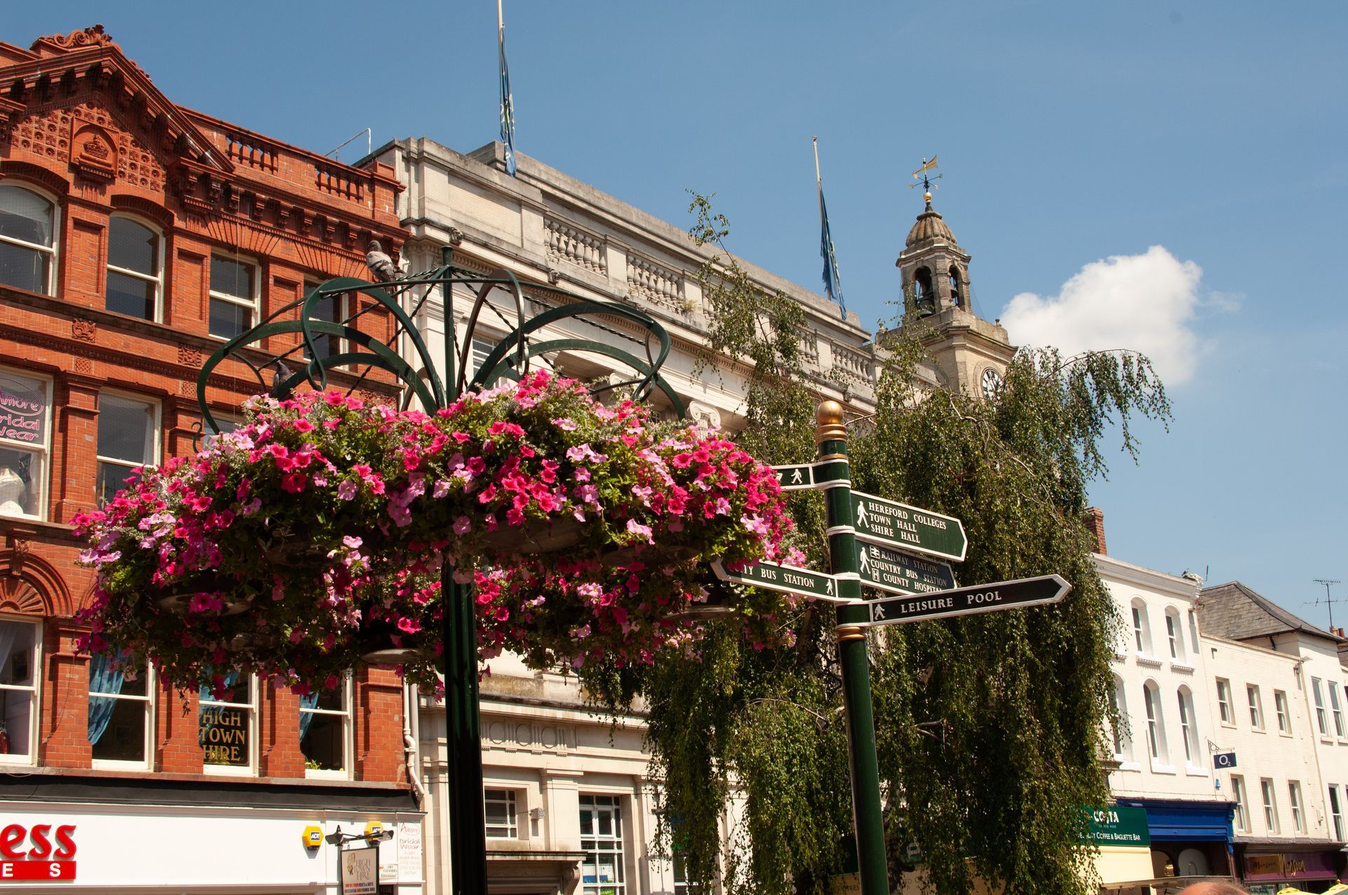 View of civic and commercial properties in Hereford town centre. Summer flower baskets hang from display stands with a signage post displaying panels with local information and directions.