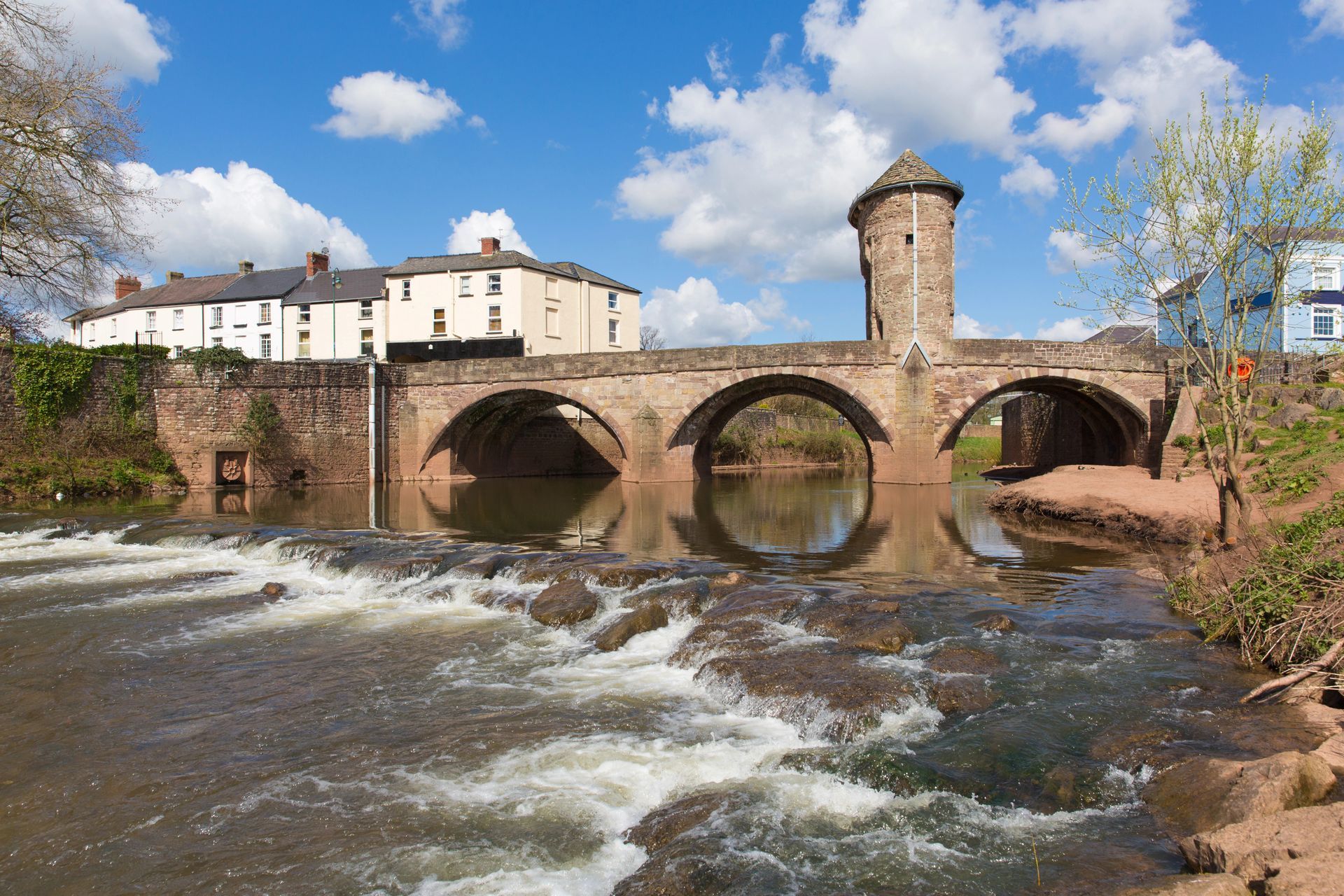 View of the historic 12th Century Monnow Bridge over the River Monnow in Monmouth, with water flowing over a small stone weir.