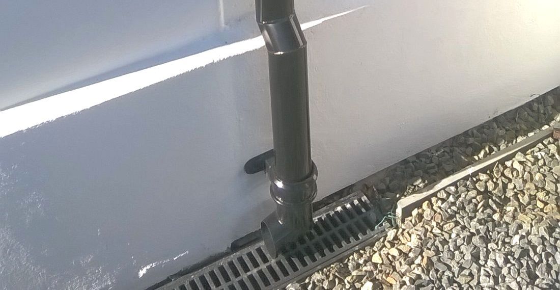a black aluminium cast gutter lays beneath a black drain pipe on a gravel base.