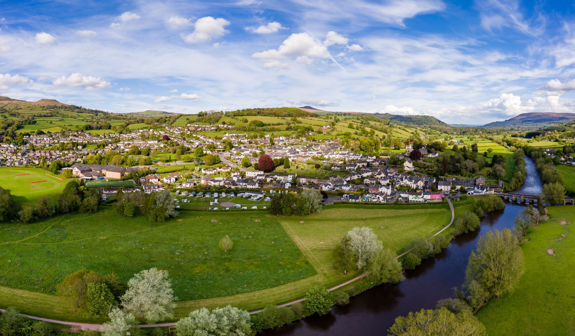 Ariel view of the pretty town of Crickhowell, Powys, Wales with the River Usk in the foreground. The town is surrounded by rolling hills with trees and mountains in the far distance.