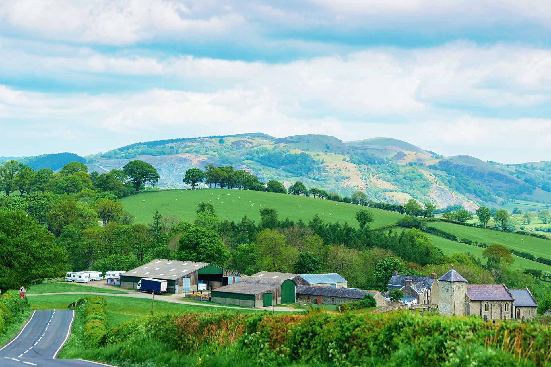 View of a rural farm and buildings in North Powys, Wales. Mountains extend up behind providing a dramatic backdrop. A winding road is positioned to the left of the farm.