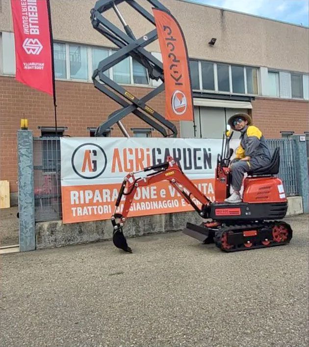 Persona che aziona un piccolo escavatore davanti a uno striscione con la scritta 