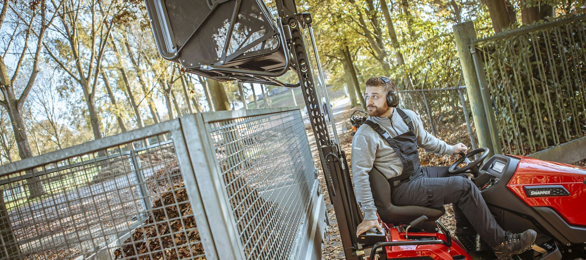 Un uomo su un trattore rosso falcia l'erba vicino a una recinzione metallica in una zona boscosa. Indossa protezioni acustiche.