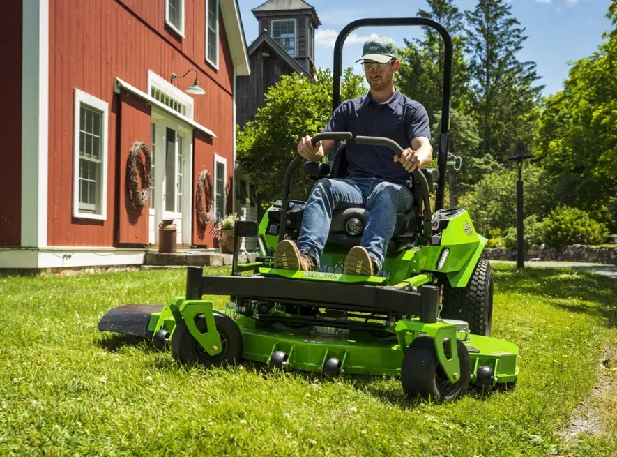 Un uomo su un tosaerba verde taglia l'erba vicino a un edificio rosso in una giornata di sole.