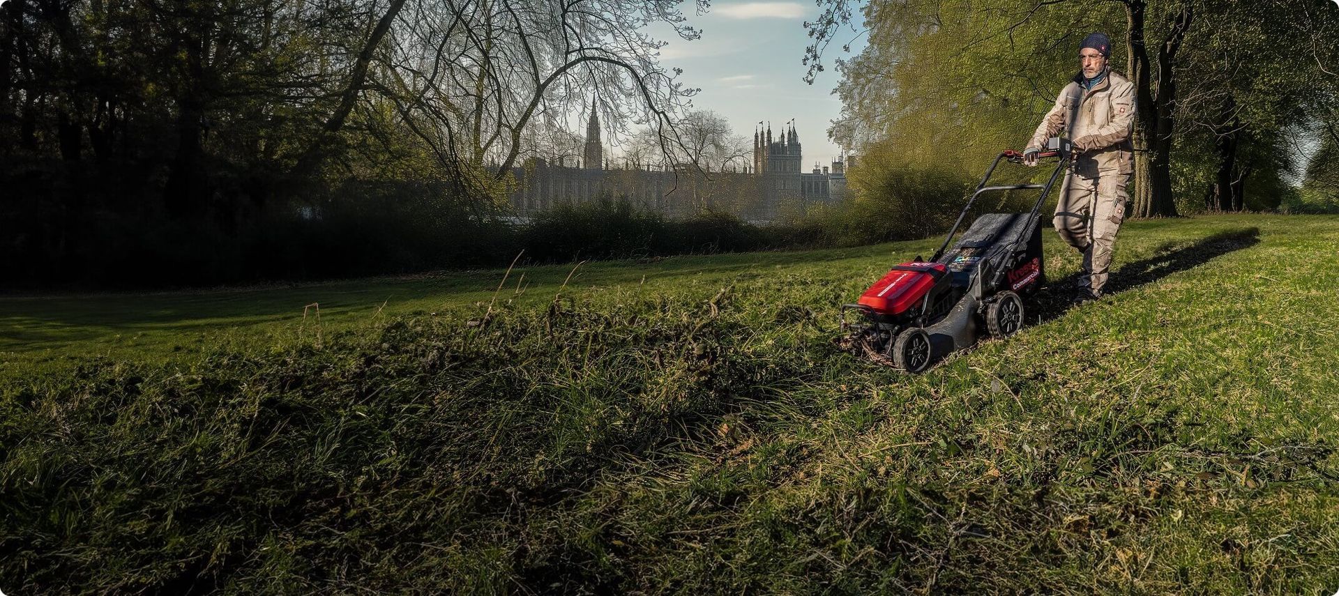 Persona che taglia l'erba con un tosaerba rosso; sullo sfondo alberi e un edificio in lontananza.