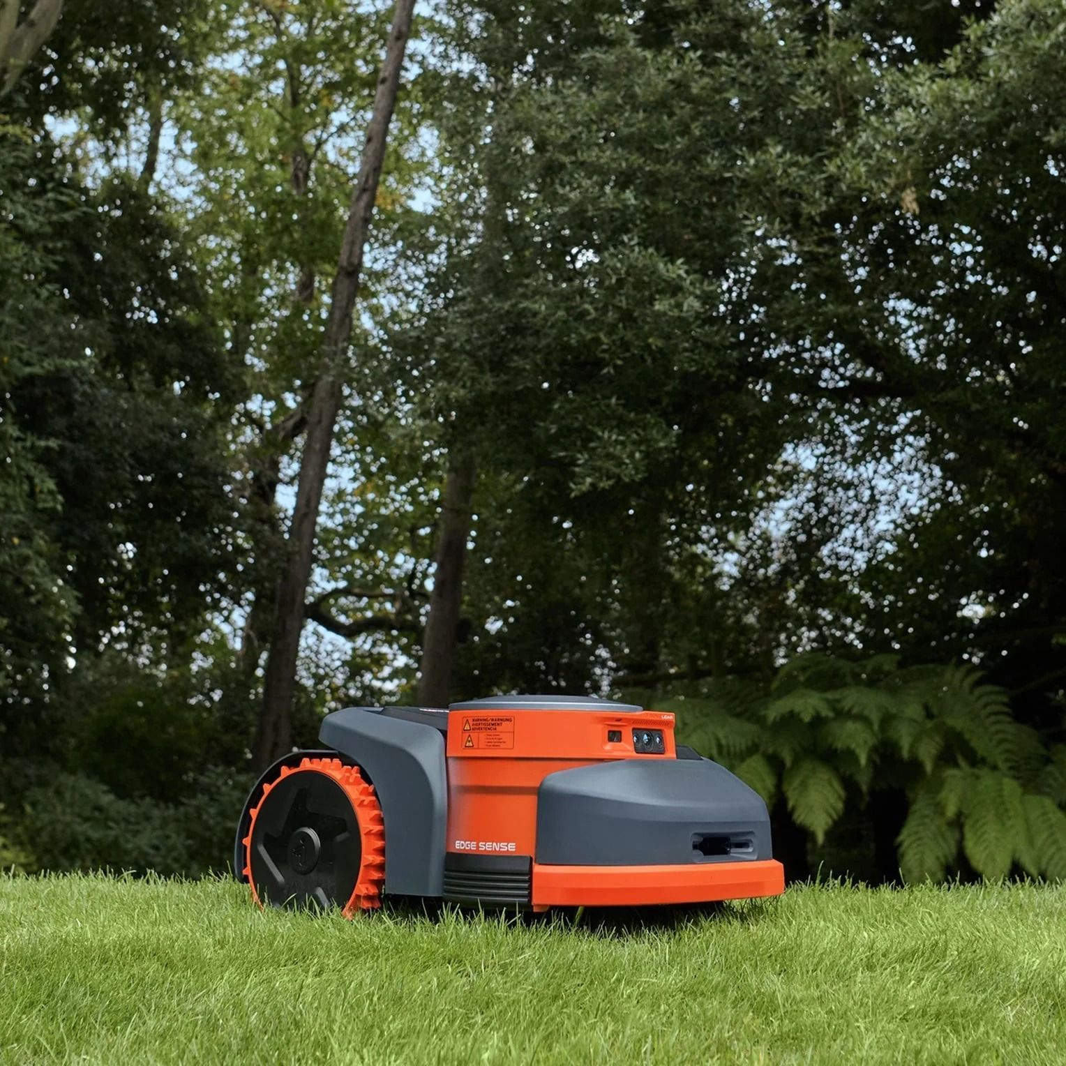 Tosaerba robotizzato sull'erba; design arancione e grigio; alberi sullo sfondo.