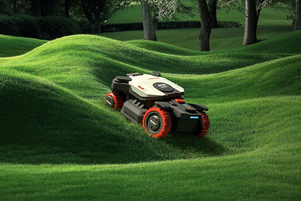 Tosaerba robotizzato su un prato verde con una fila di alberi sullo sfondo.