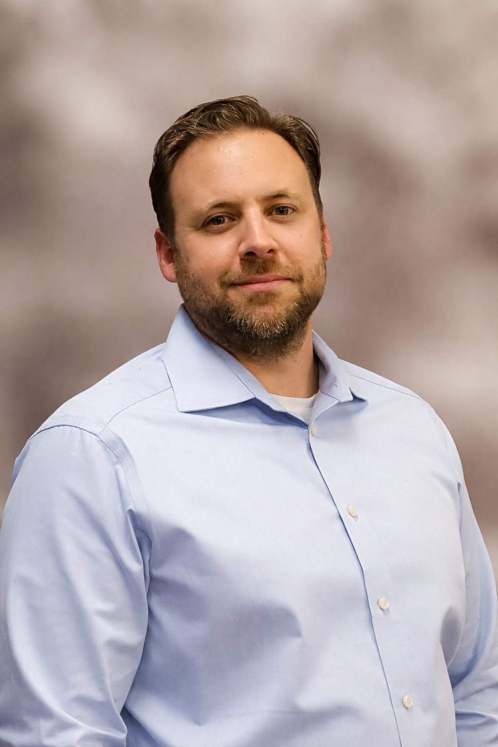 Een persoon met kort bruin haar en een baard, gekleed in een lichtblauw overhemd, glimlacht tegen een neutrale achtergrond.