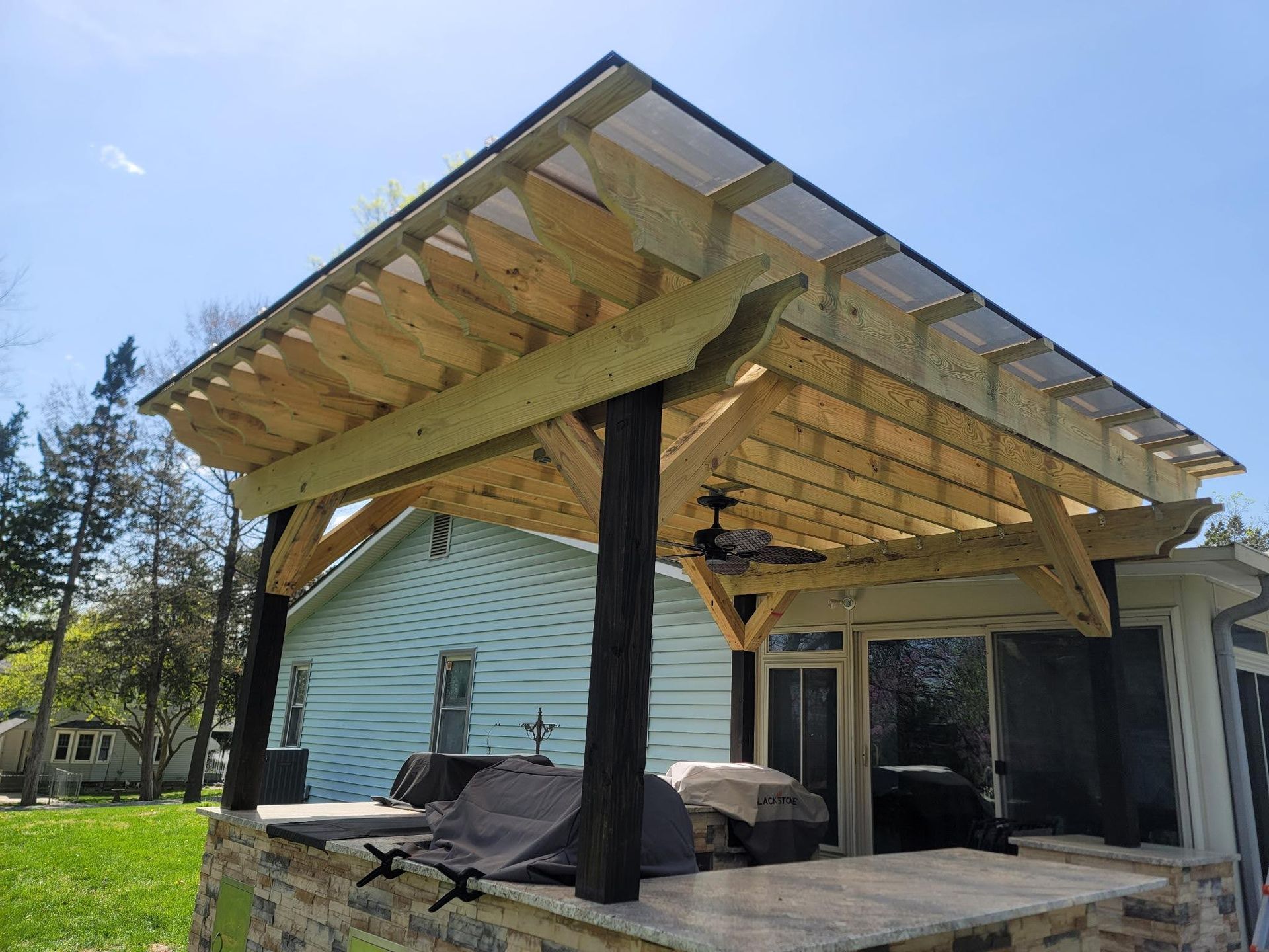 A wooden pergola with a ceiling fan is sitting in front of a house.