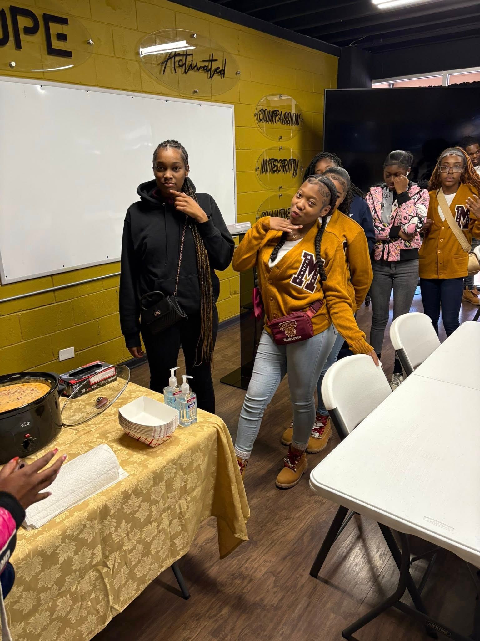 Group of people standing in a room with a table, whiteboard, and food. One woman poses.