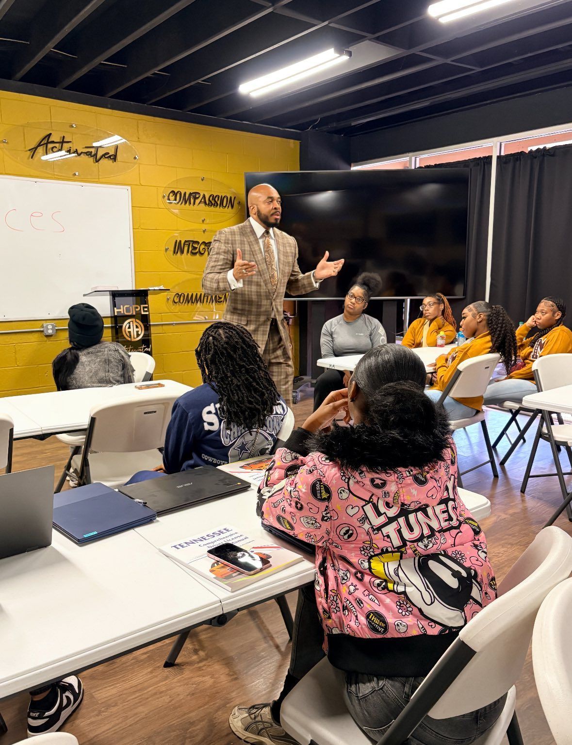 A man in a suit leads a class, gesturing with his hands. Students seated at desks, some with laptops. Yellow and black room.