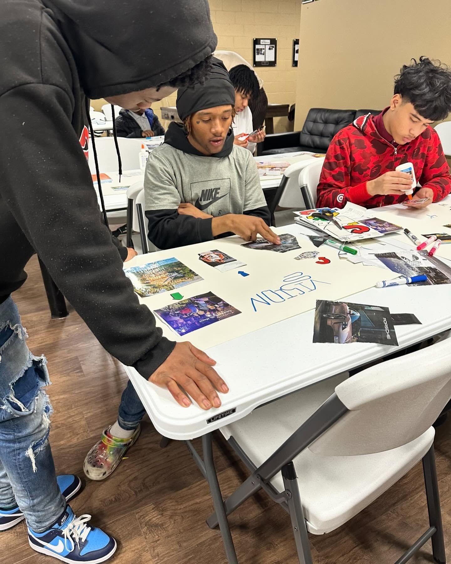 Three people at a white table look at pictures, a board. Someone wearing a black hoodie points to the board. Indoors.